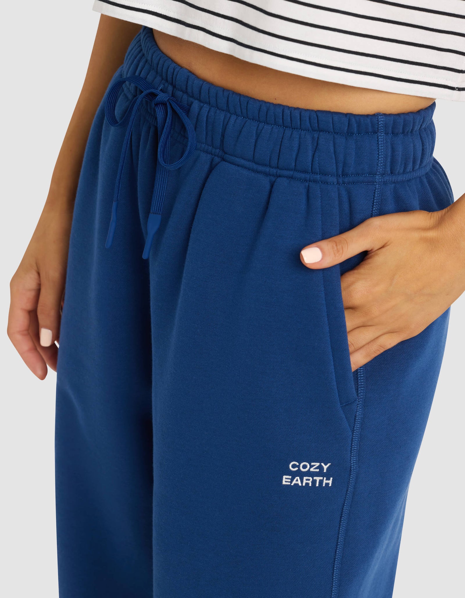 A woman wears Cozy Earth's Women's Cityscape 2.0 Sweatpant in blue, featuring the brand name on the pocket, paired with a white striped crop top and one hand in her pocket.