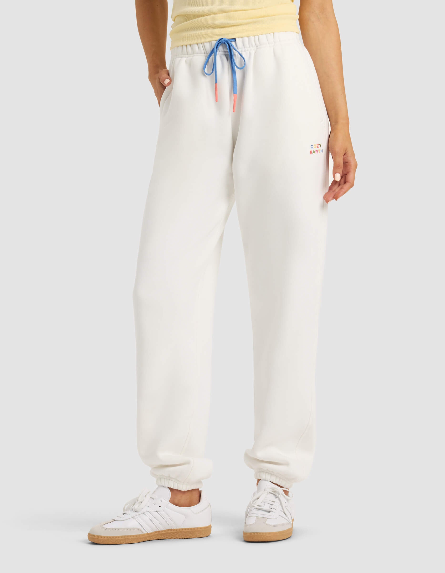 A person models Cozy Earth's Women's Cityscape 2.0 Sweatpant in white, paired with a yellow top and white sneakers, hands in pockets, against a plain background. 
