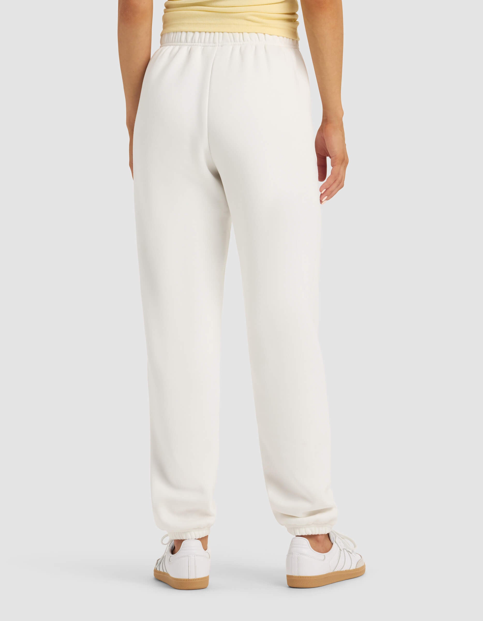 A person is seen from behind wearing the Cozy Earth Women's Cityscape 2.0 Sweatpant in white, paired with a light yellow top and white sneakers, standing against a plain light gray background.