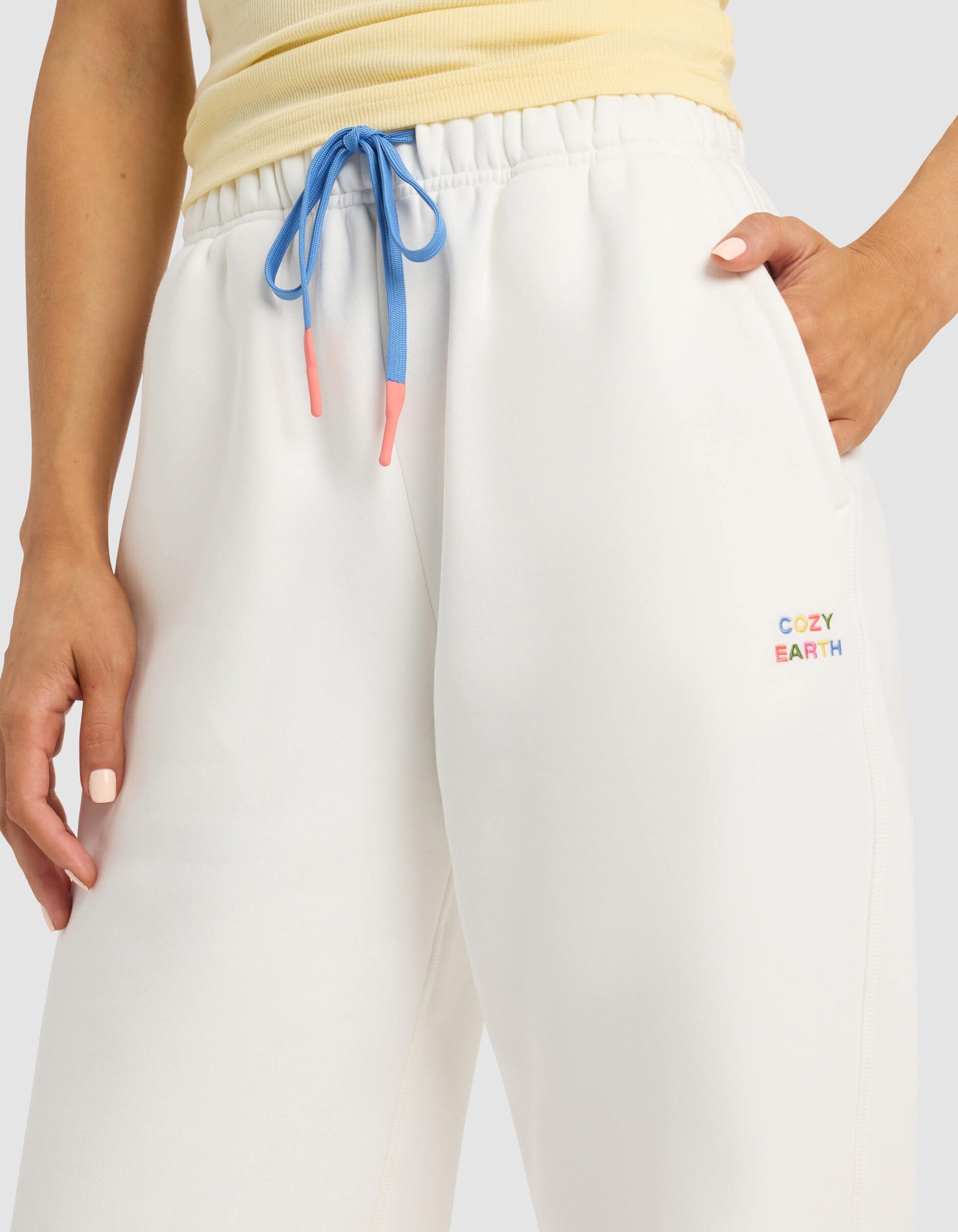 A person wears Cozy Earth Women's Cityscape 2.0 Sweatpant in white with blue drawstrings and a multicolored logo, standing with one hand in their pocket. The top of a light yellow shirt is also visible.