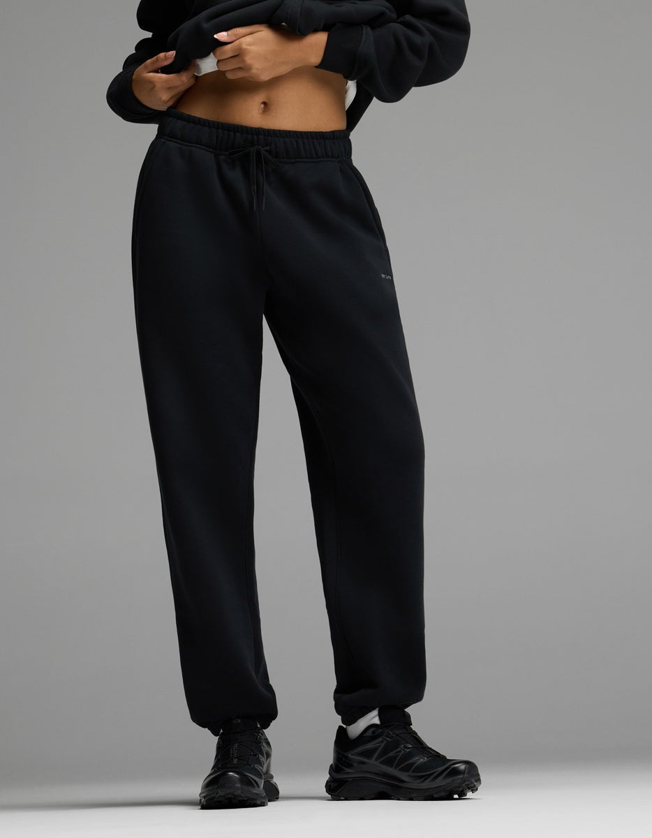 A person wearing Cozy Earth’s Women’s CityScape Sweatpant in black, paired with black sneakers and a cropped black hoodie, stands against a gray background with hands near the waistband.