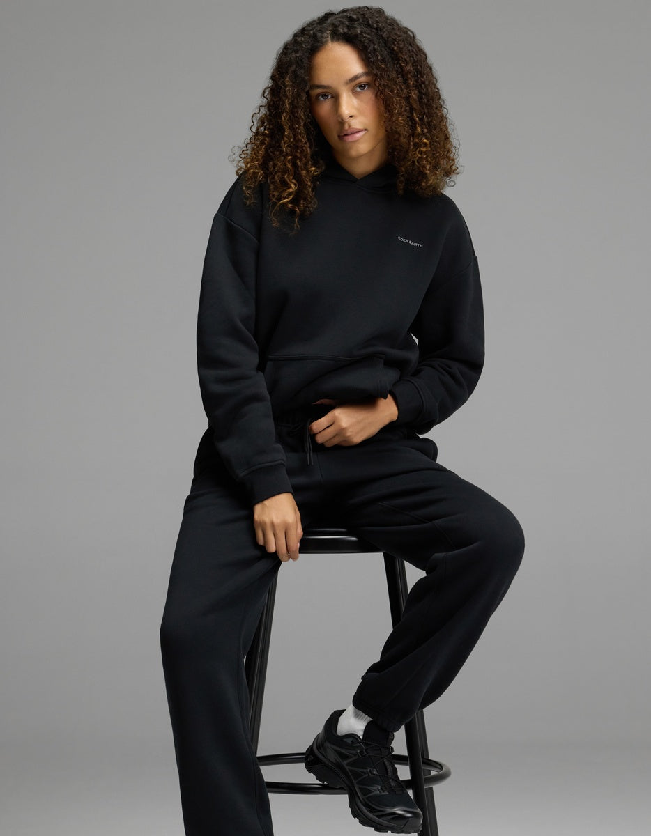 A person with curly hair wearing a black hoodie, Cozy Earth Women’s CityScape Sweatpant in black, and black sneakers sits on a black stool against a plain gray background.