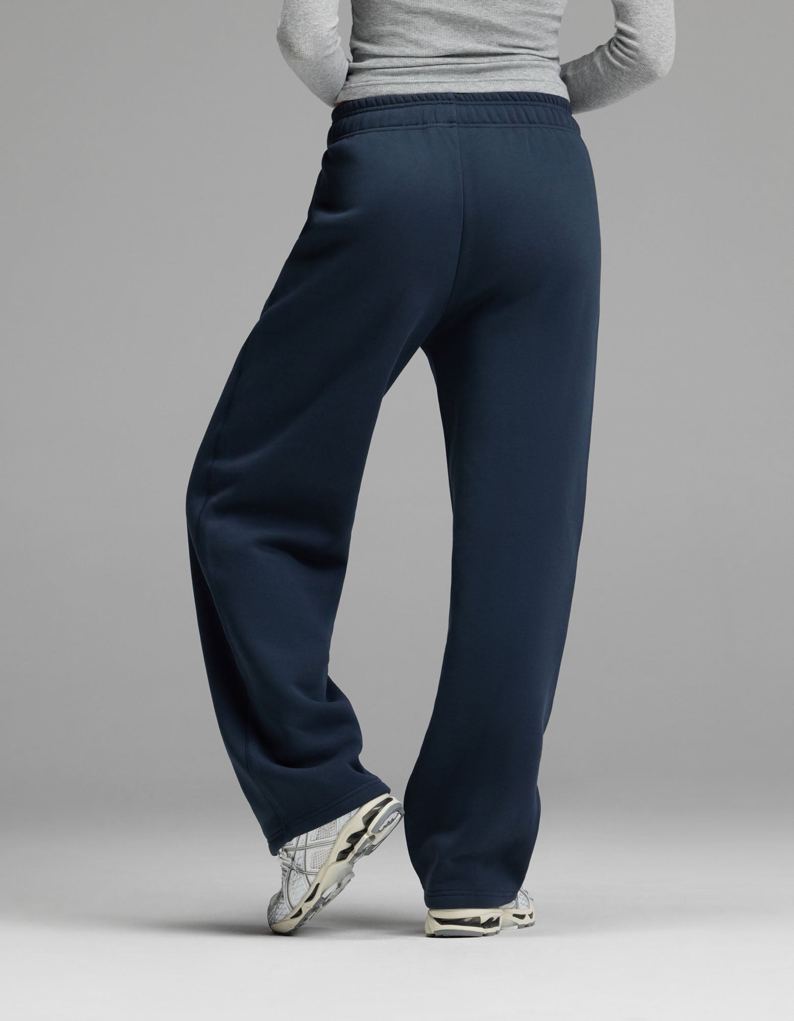 A person wearing a gray long-sleeve shirt, Cozy Earth Women’s Cityscape Wide Leg Pant in navy blue, and white sneakers stands with legs crossed, facing away from the camera against a plain gray background.