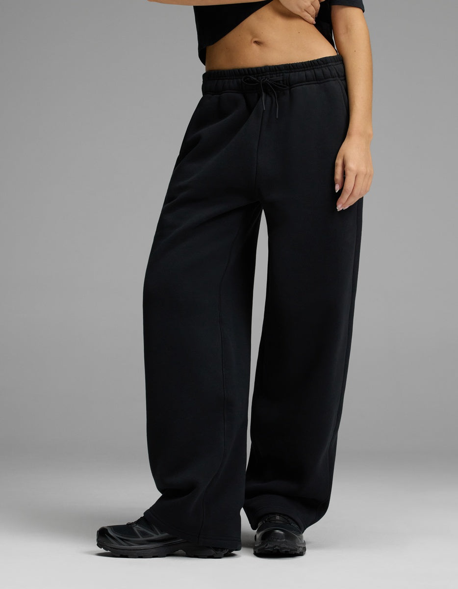 A person wearing the Cozy Earth Women’s CityScape Wide Leg Pant in Jet Black stands against a plain gray background, paired with a black cropped top and black sneakers. Their midriff and one hand are visible.