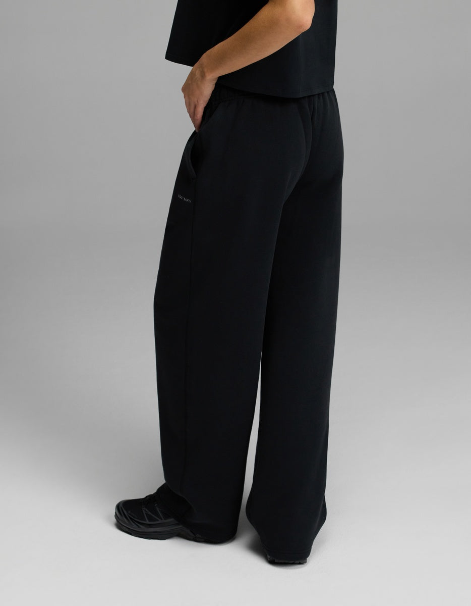 A person stands with one hand in their pocket, wearing Cozy Earth’s Women’s CityScape Wide Leg Pant in black, paired with a black top and black sneakers against a plain gray background.