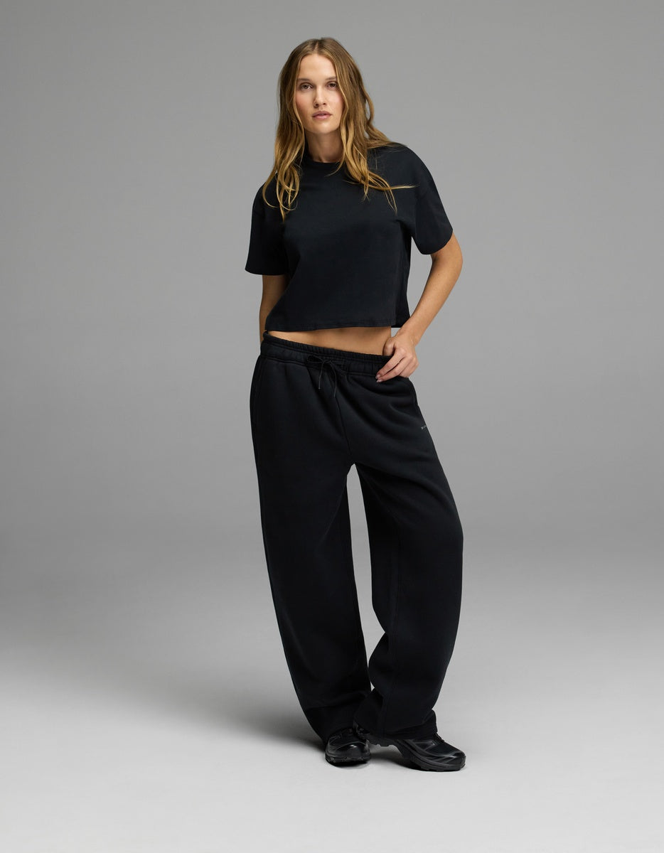 A woman stands against a plain gray background, wearing a black short-sleeve crop top, black sneakers, and Cozy Earth’s Women’s CityScape Wide Leg Pant. Her left hand rests in her pocket as she looks confidently at the camera.