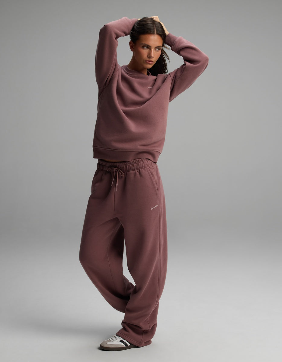 A person wearing a dusty mauve Women’s CityScape Crewneck sweatshirt and matching sweatpants with a drawstring waist, paired with grey and black sneakers.