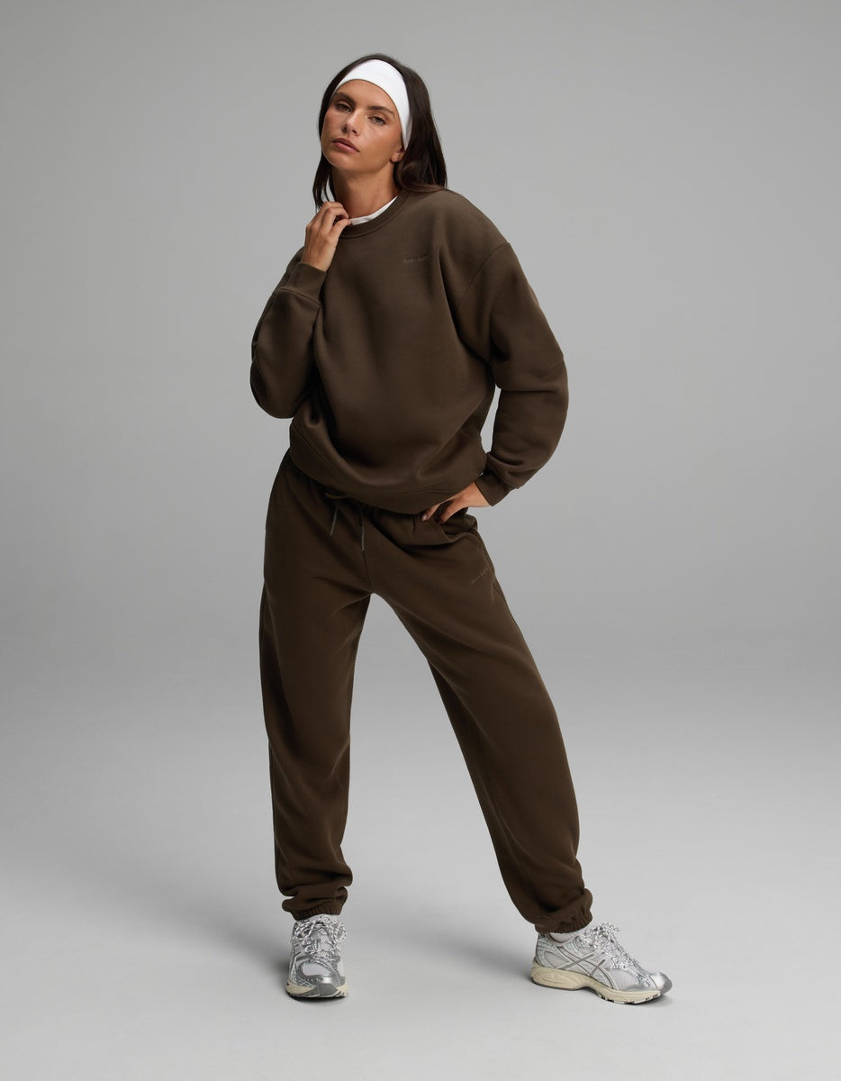 A person wearing a dark brown Cozy Earth Women’s Cityscape Crewneck & Sweatpant Set with a white headband and metallic athletic shoes.