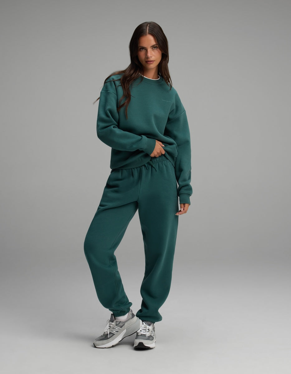 A person wearing a deep green Cozy Earth Women’s Cityscape Crewneck & Sweatpant Set with grey sneakers.