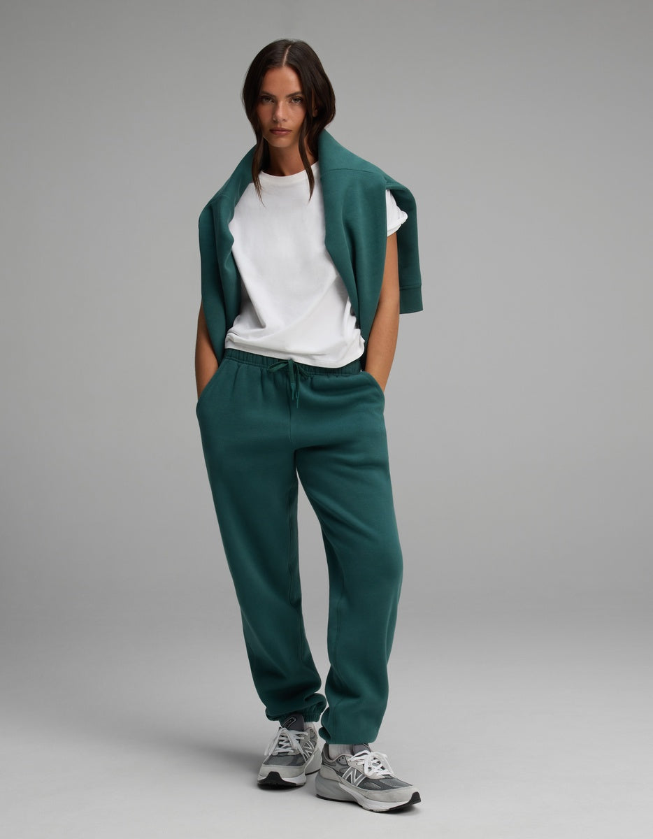 A model wears a white t-shirt and the Cozy Earth Women’s Cityscape Crewneck & Sweatpant Set, featuring dark green sweatpants with a drawstring waist and a matching green sweatshirt draped over her shoulders.