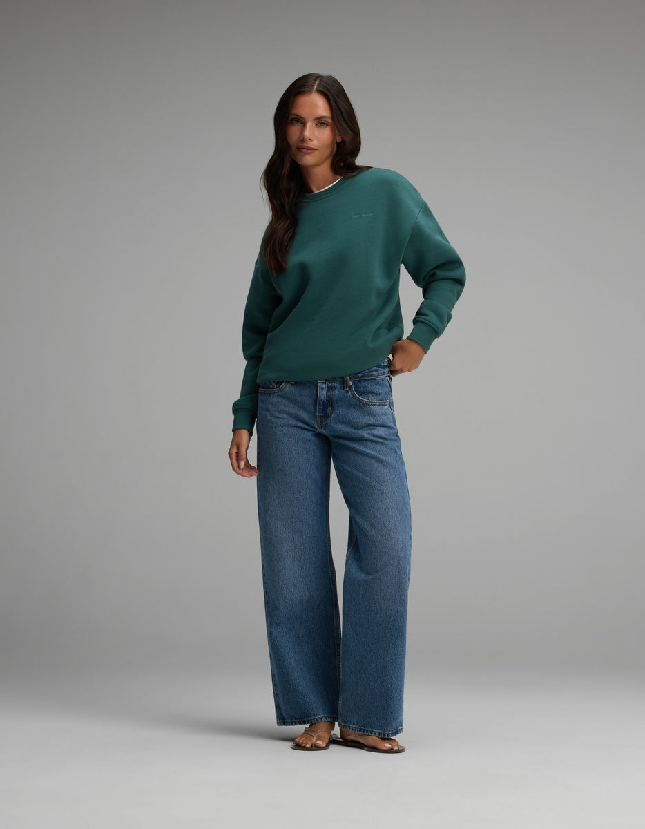 A woman wears a forest green Cozy Earth Women’s CityScape Crewneck and blue wide-leg jeans.