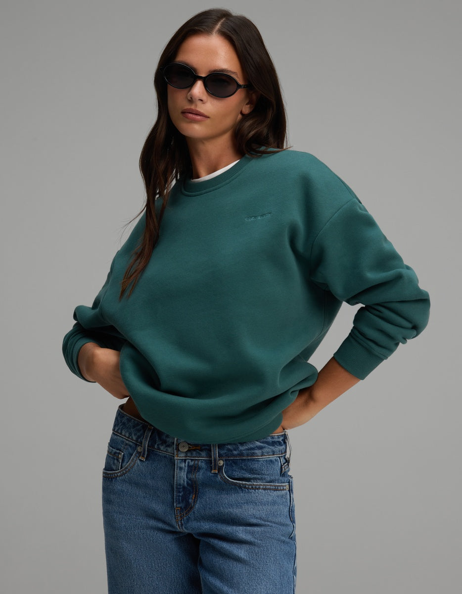 A person wearing a teal Cozy Earth Women’s CityScape Crewneck and blue jeans.
