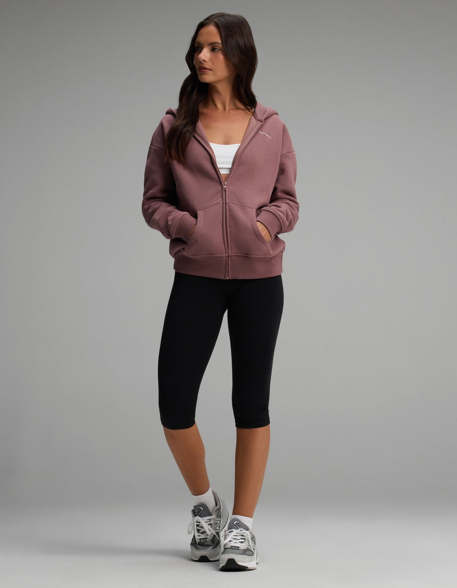 A person wearing a dusty rose Women’s CityScape Full-Zip Hoodie, white sports bra, black capri leggings, and grey athletic shoes.