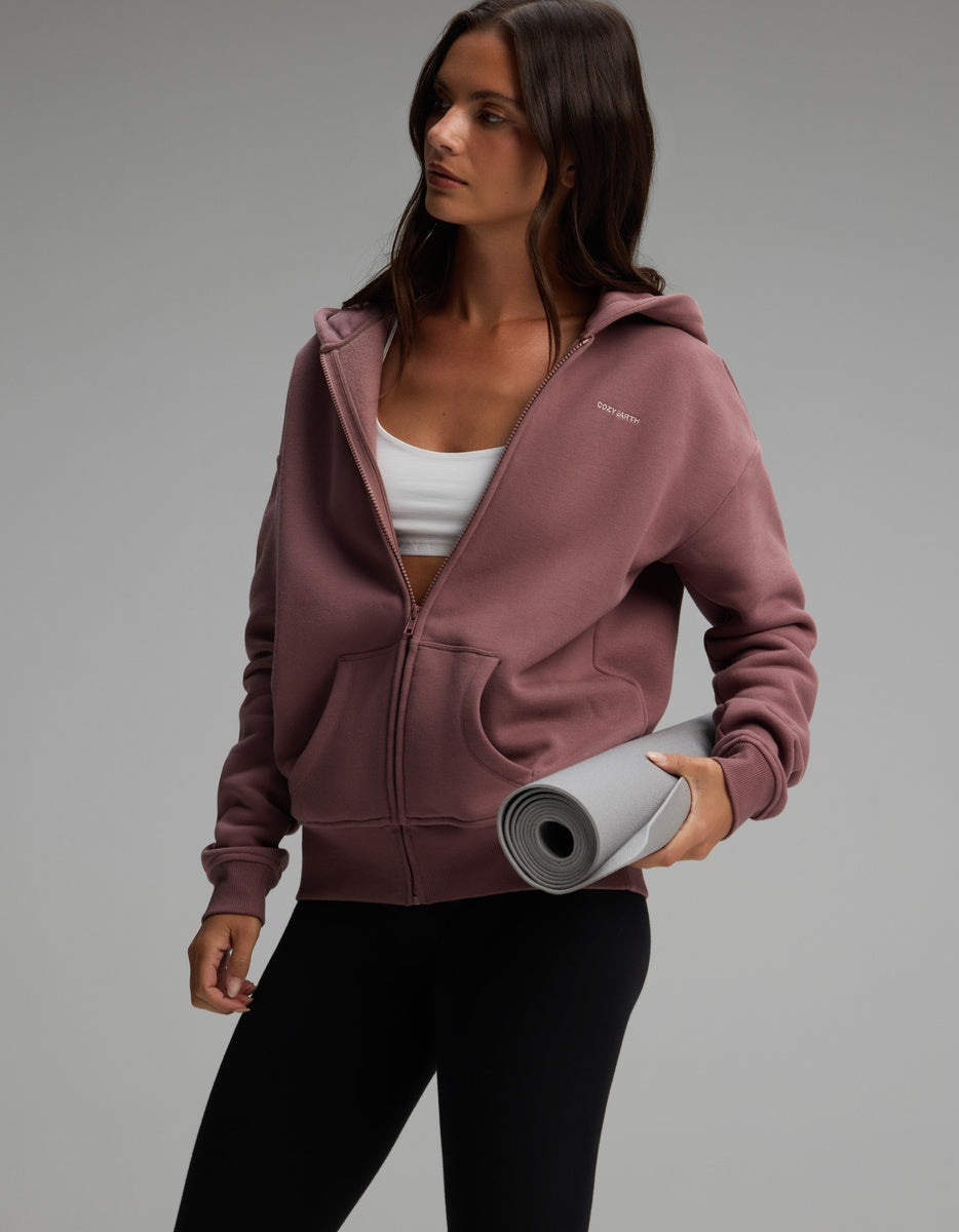 A woman wears a mauve Women’s CityScape Full-Zip Hoodie over a white sports bra, holding a grey rolled yoga mat.