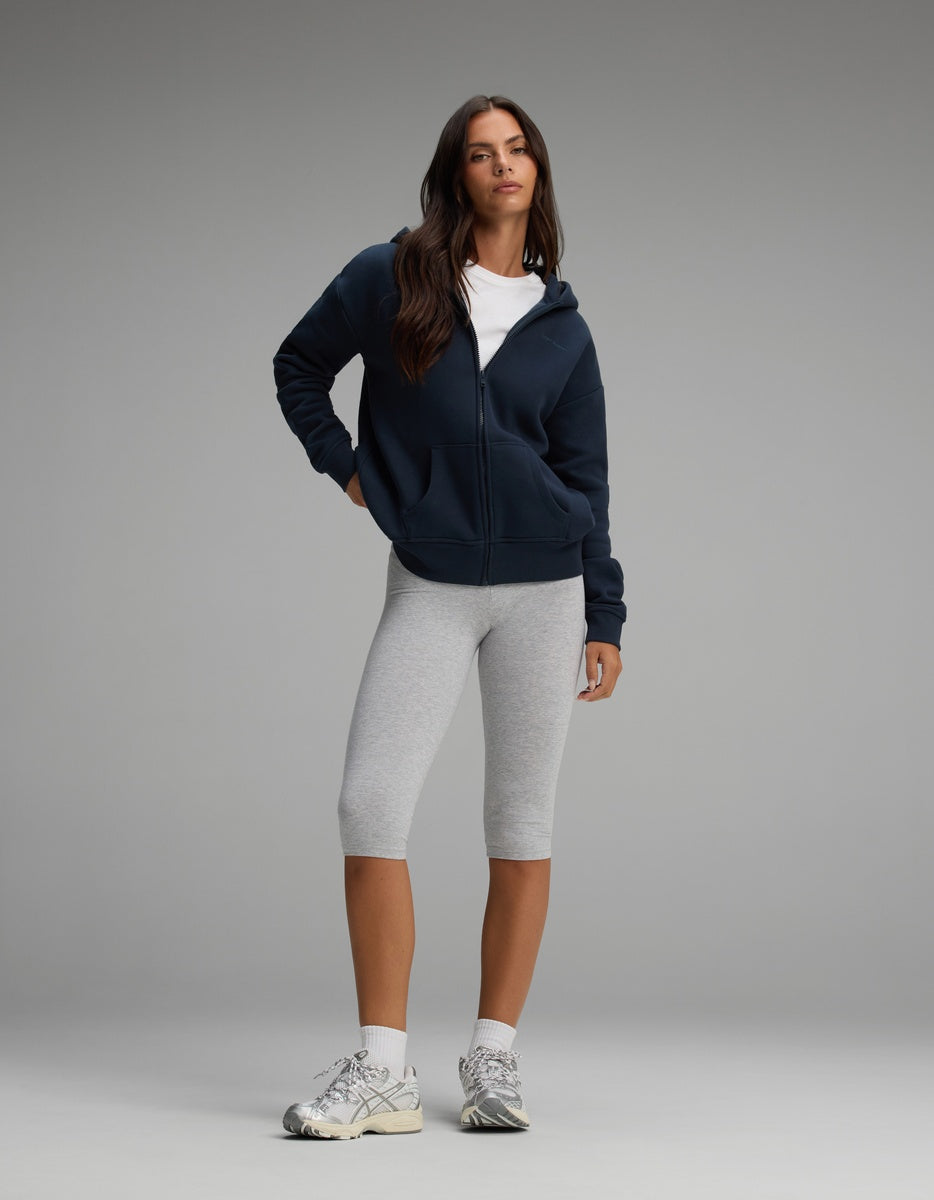 A woman wearing a dark blue Cozy Earth Women’s CityScape Full-Zip Hoodie over a white t-shirt, light gray cropped leggings, white socks, and sneakers.