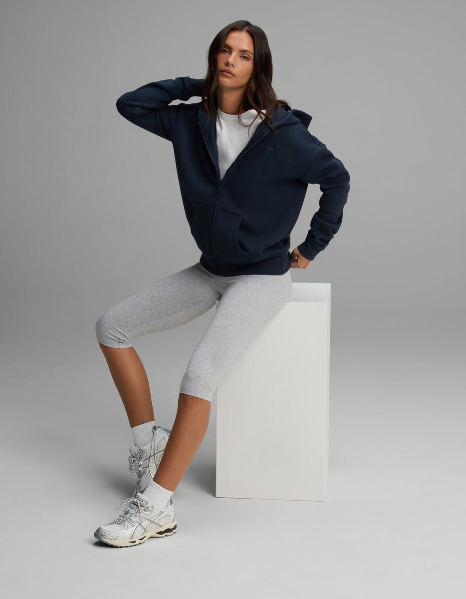 A woman wearing a navy blue Cozy Earth Women’s CityScape Full-Zip Hoodie, white t-shirt, and gray cropped leggings sits on a white cube.