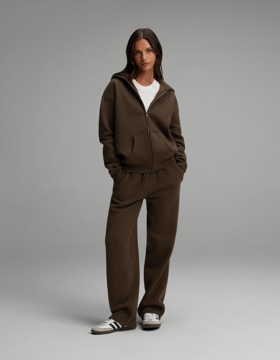 A woman wearing a dark brown Cozy Earth Women’s CityScape Full-Zip Hoodie and matching wide-leg sweatpants over a white crew-neck shirt.