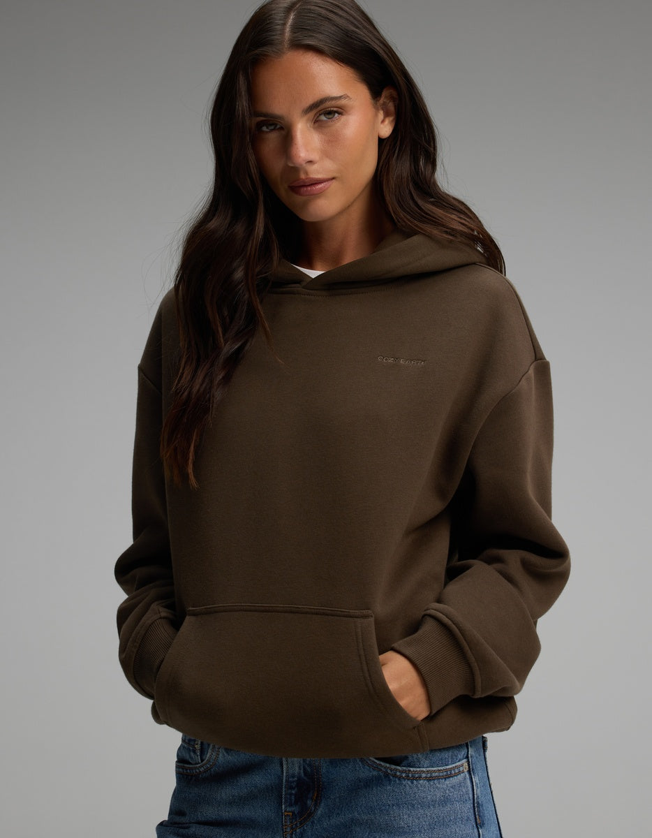 A woman wearing a brown Cozy Earth Women’s CityScape Hoodie with a front kangaroo pocket and jeans.