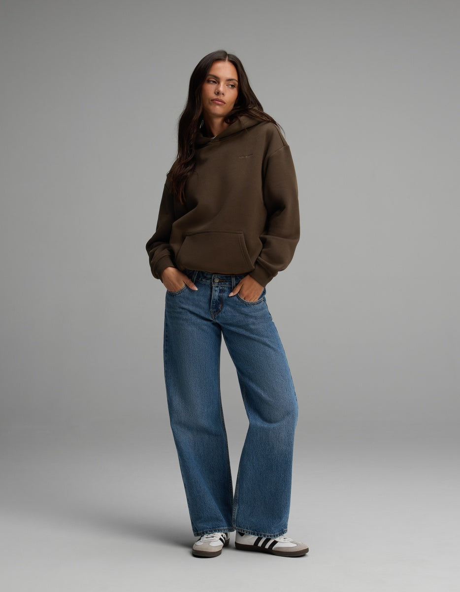 A person wearing a dark brown Cozy Earth Women’s CityScape Hoodie, blue jeans, and sneakers with their hands in the front pocket.