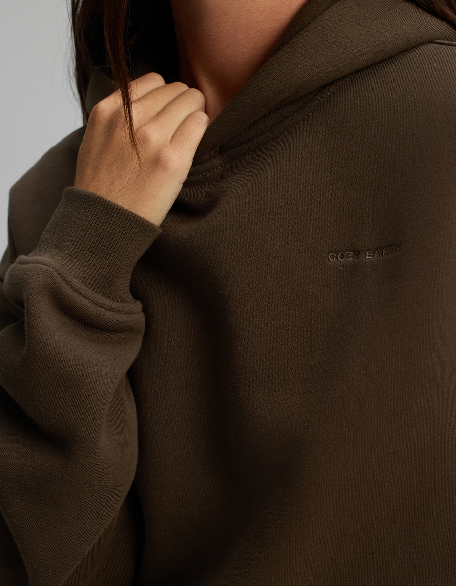 Close-up of a person wearing a dark brown Cozy Earth Women’s CityScape Hoodie with the brand name embroidered on the chest.
