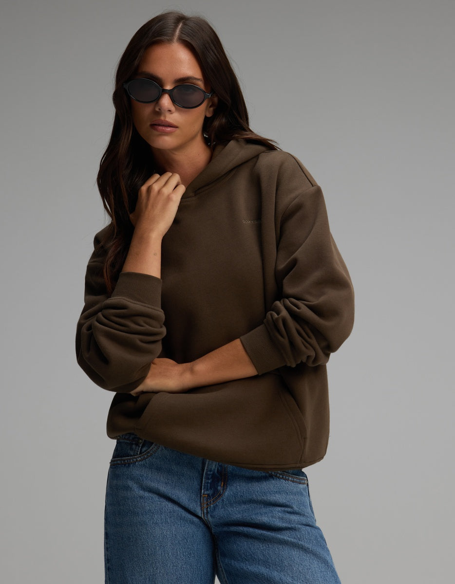 A person wearing a dark olive Women’s CityScape Hoodie and blue jeans.