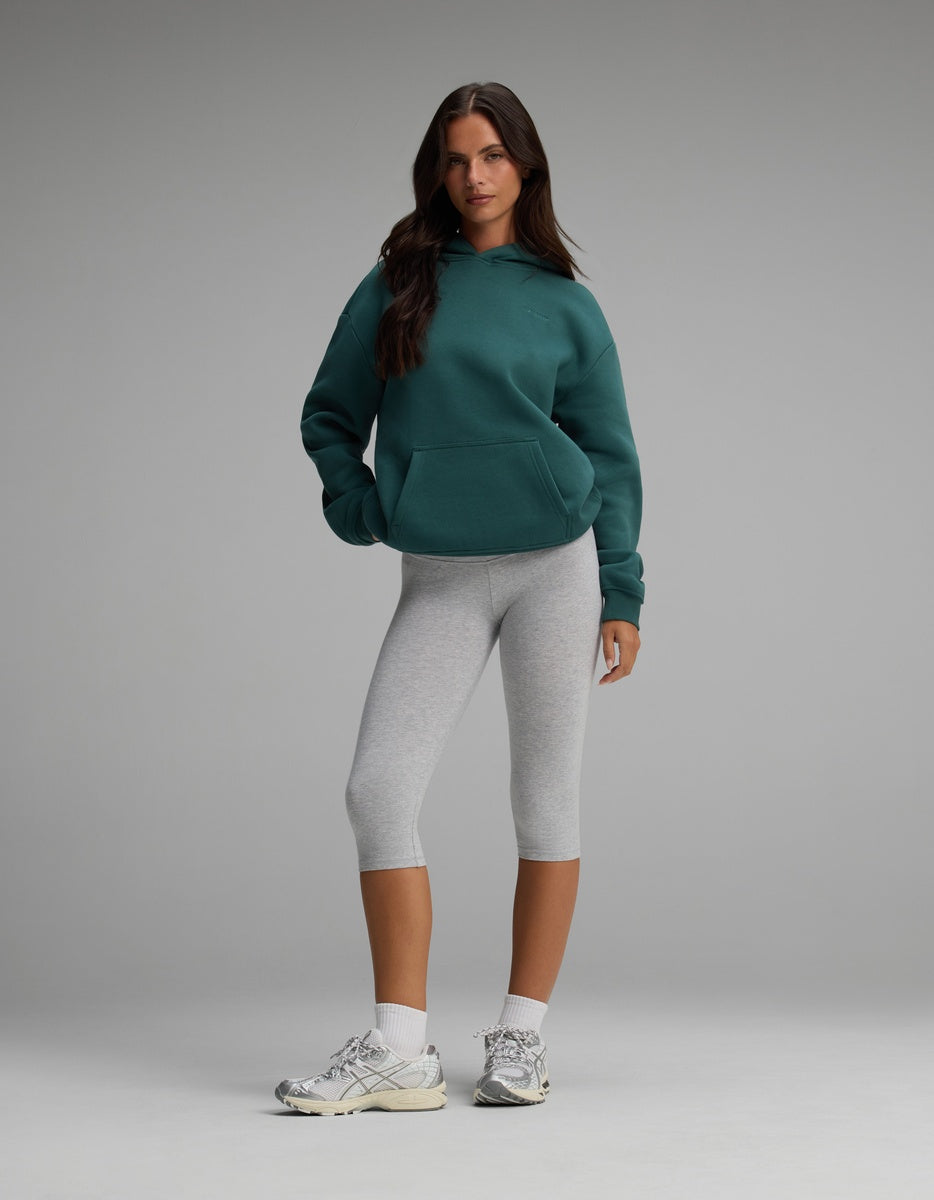 A person wearing a dark teal Cozy Earth Women’s CityScape Hoodie, grey cropped leggings, white socks, and athletic sneakers.
