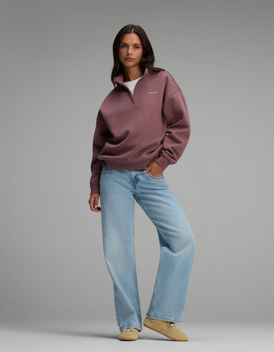 A woman wearing a mauve Cozy Earth Women’s CityScape Quarter Zip, light wash wide-leg jeans, and tan sneakers.