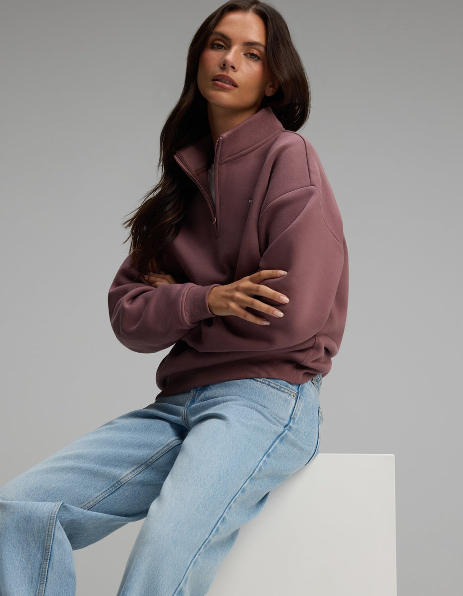 A person wearing a dusty rose-colored Cozy Earth Women’s CityScape Quarter Zip and light-wash blue jeans sitting with arms crossed.