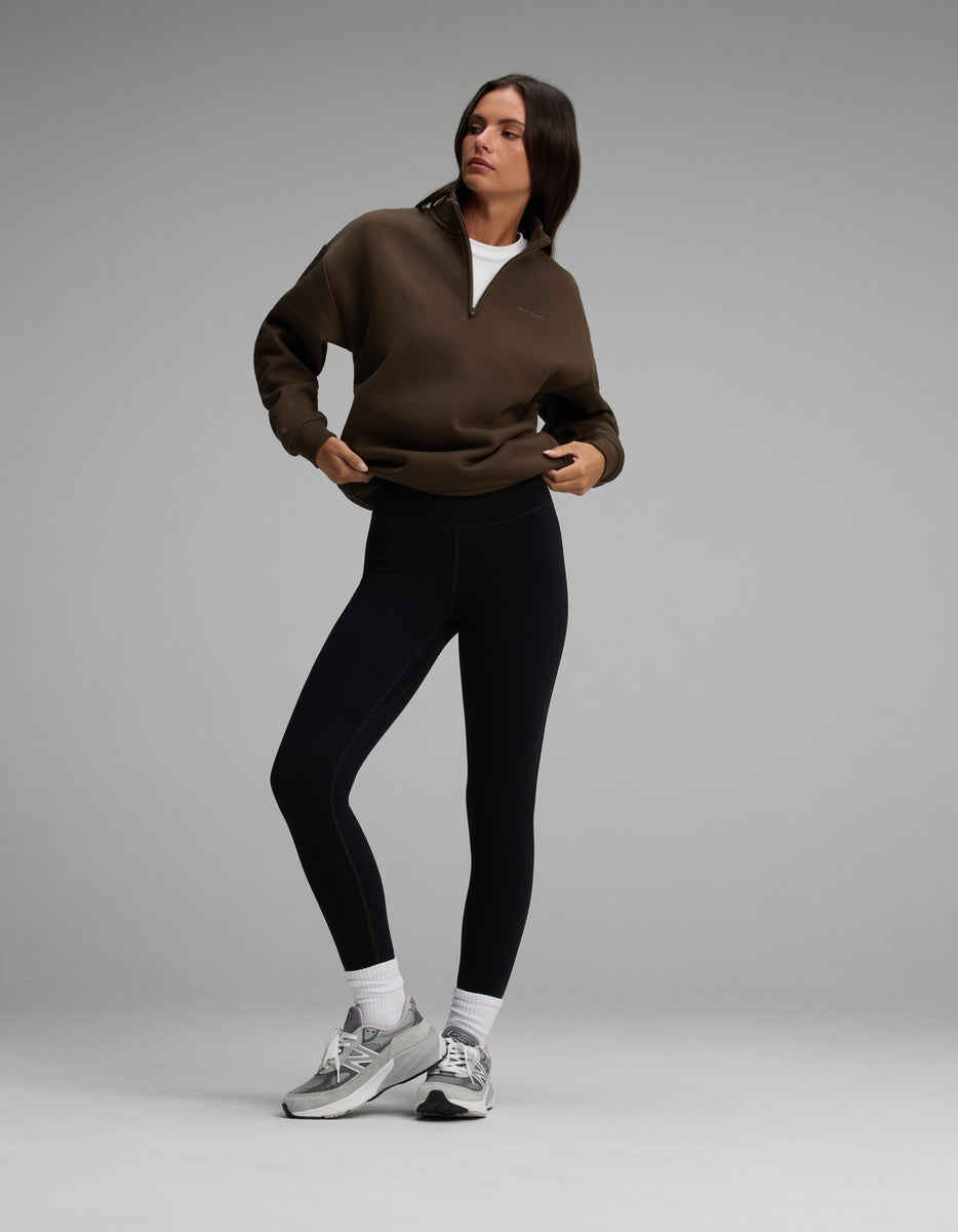 A woman wearing a dark brown Cozy Earth Women’s CityScape Quarter Zip over a white shirt, black leggings, and grey athletic sneakers.