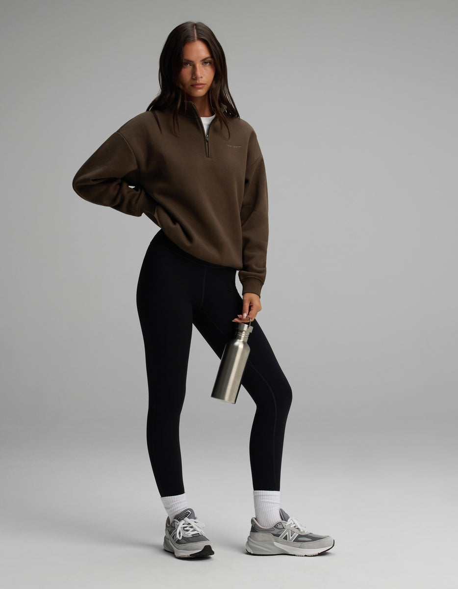A woman wearing a brown Cozy Earth Women’s CityScape Quarter Zip, black leggings, white socks, and athletic sneakers, holding a metal water bottle.