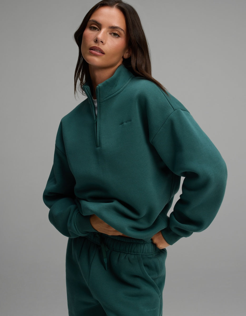 A woman wearing a dark green Cozy Earth Women’s CityScape Quarter Zip with a high collar and matching drawstring sweatpants.