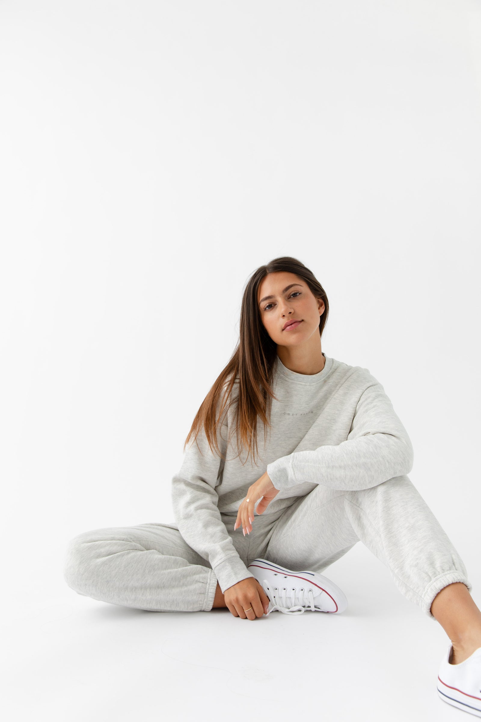 A woman with long brown hair relaxes against a white background, wearing the Women's CityScape Crewneck & Sweatpant Set by Cozy Earth and white sneakers, with one arm resting casually on her knee.|Color:Heather Grey