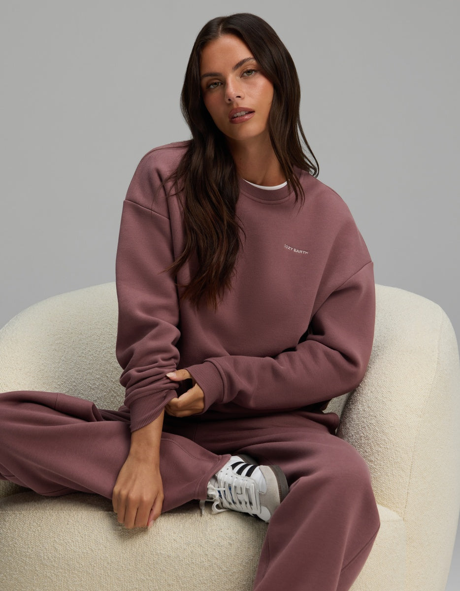 A person wearing a mauve Cozy Earth Women’s Cityscape Crewneck & Wide Leg Pant Set, seated in a white armchair.