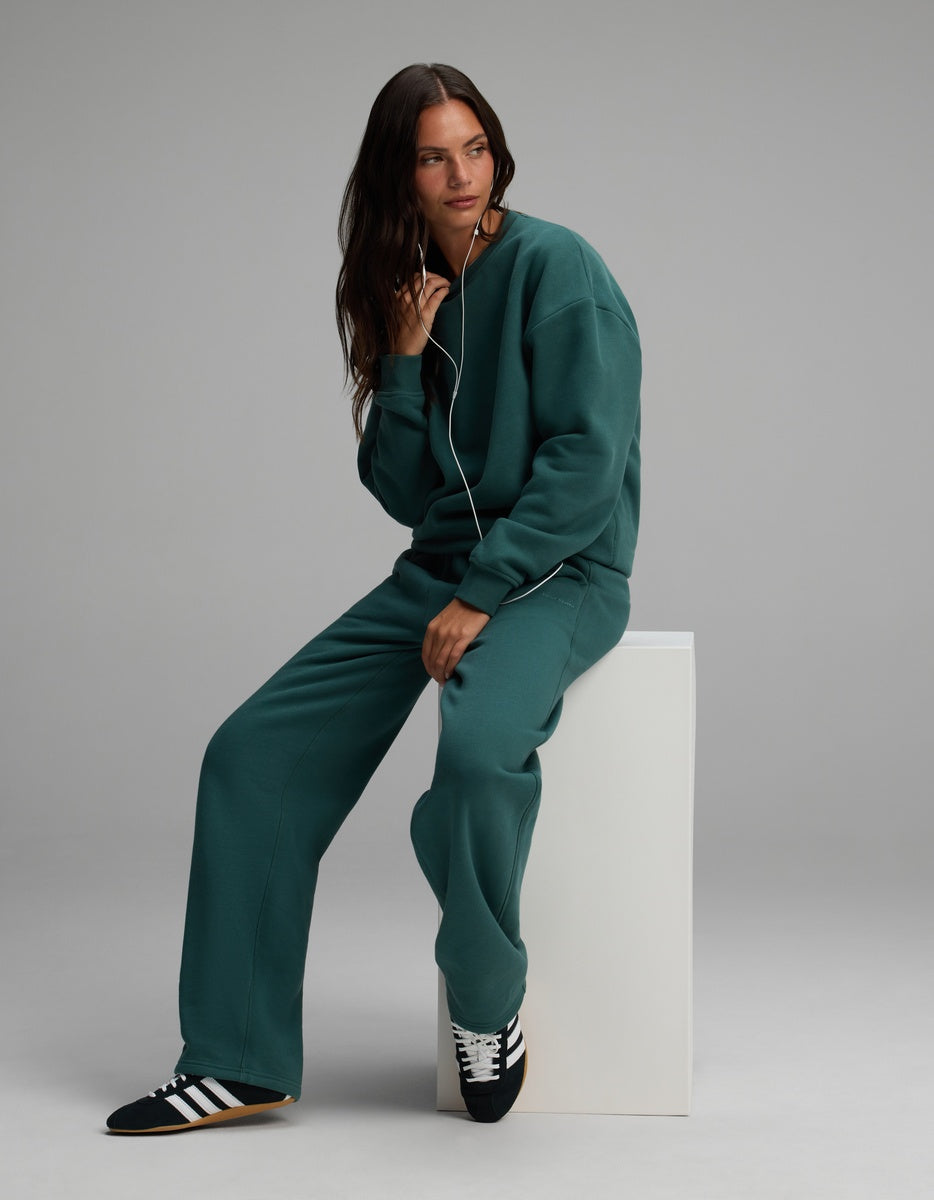 A woman wearing a dark green Cozy Earth Women’s Cityscape Crewneck & Wide Leg Pant Set, sitting on a white cube.