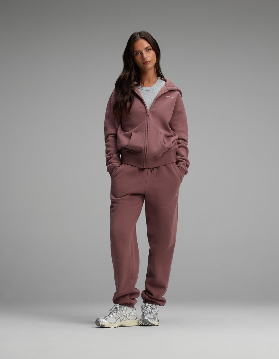 A person wears a mauve Women’s Cityscape Full-Zip Hoodie & Sweatpant Set over a grey crew-neck t-shirt, paired with white athletic sneakers.