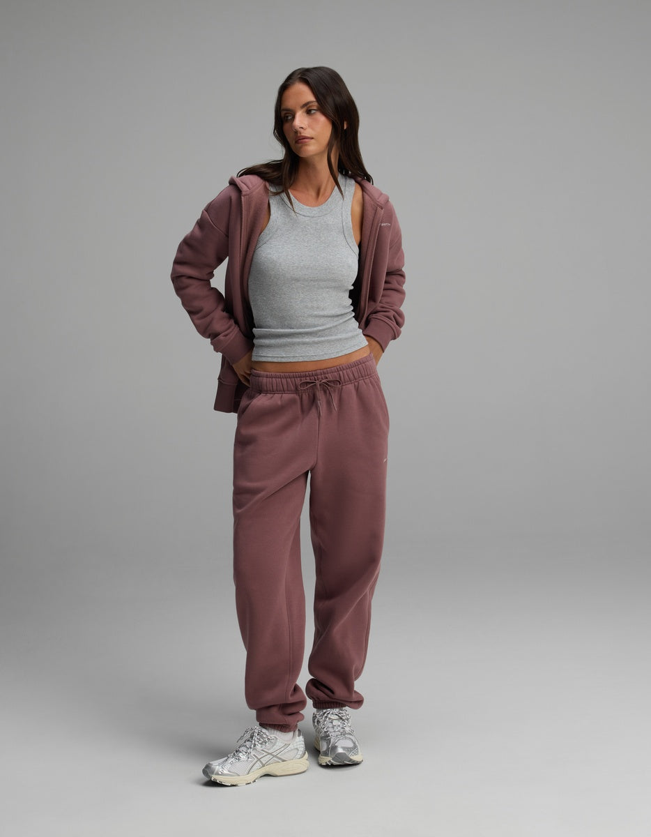 A woman wears a mauve Cozy Earth Women’s Cityscape Full-Zip Hoodie & Sweatpant Set over a grey ribbed tank top, paired with silver athletic shoes.