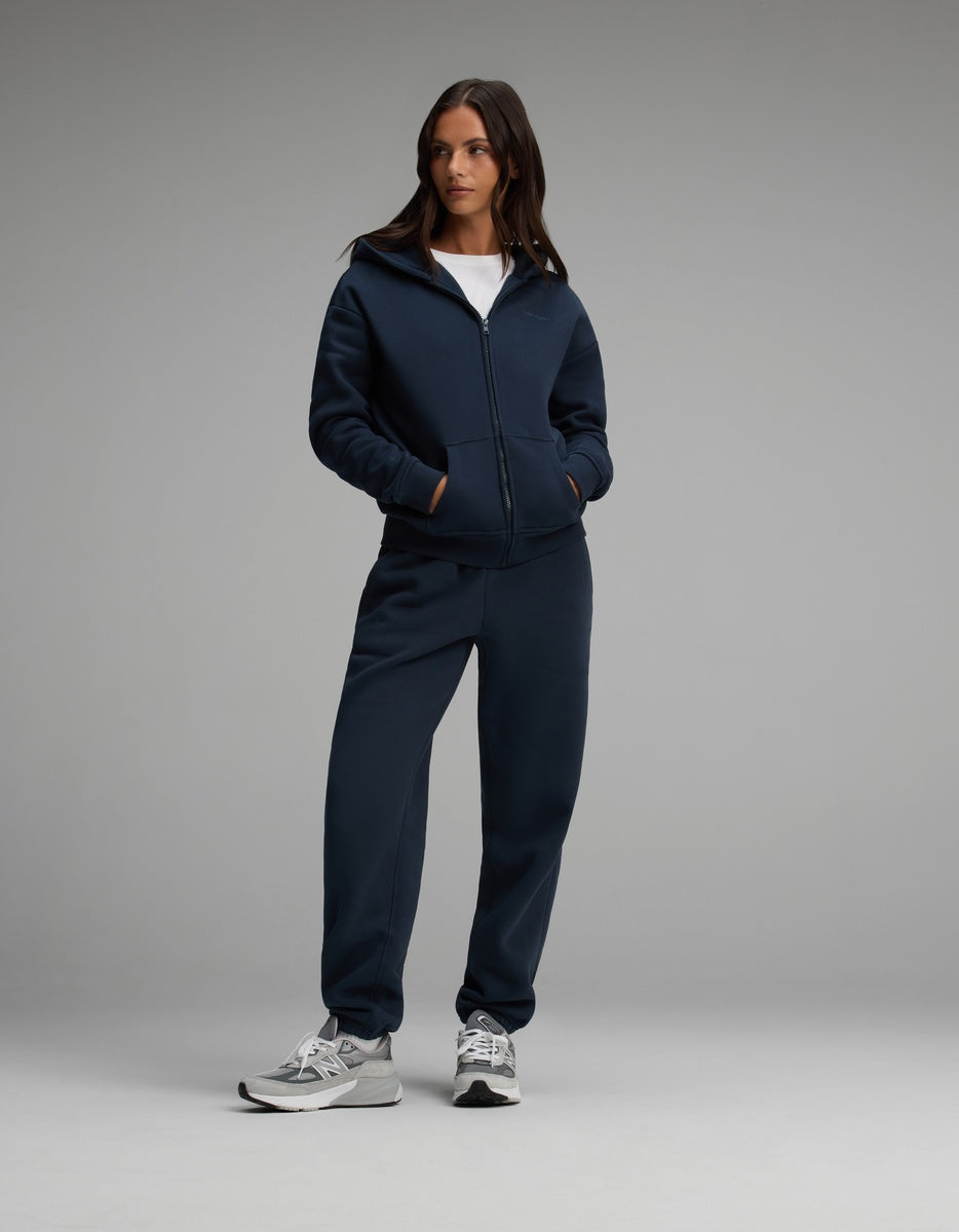 A person wearing a navy blue Women’s Cityscape Full-Zip Hoodie & Sweatpant Set with matching sweatpants and grey athletic shoes.