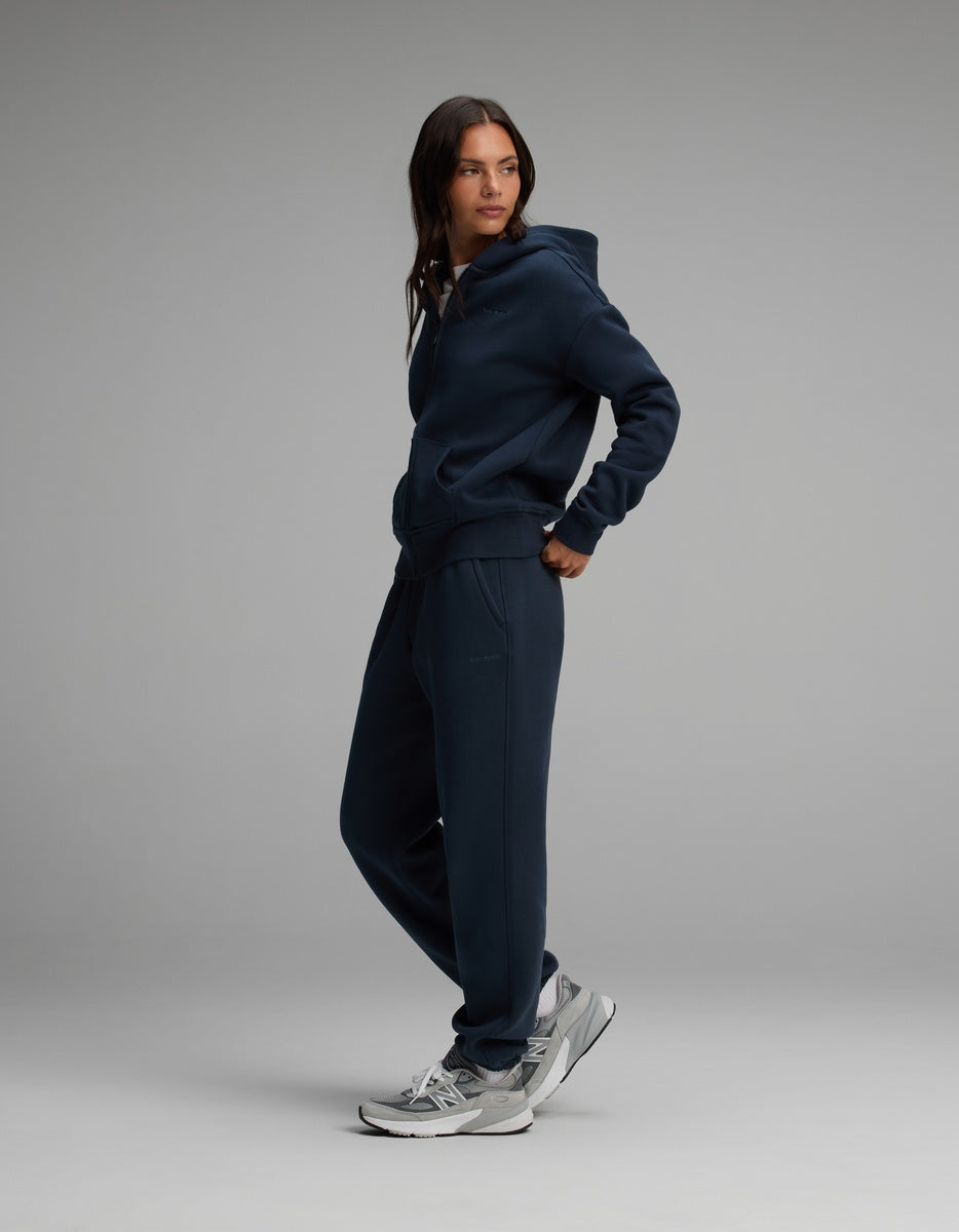 A person wearing a navy blue Cozy Earth Women’s Cityscape Full-Zip Hoodie & Sweatpant Set, paired with grey and white athletic sneakers.