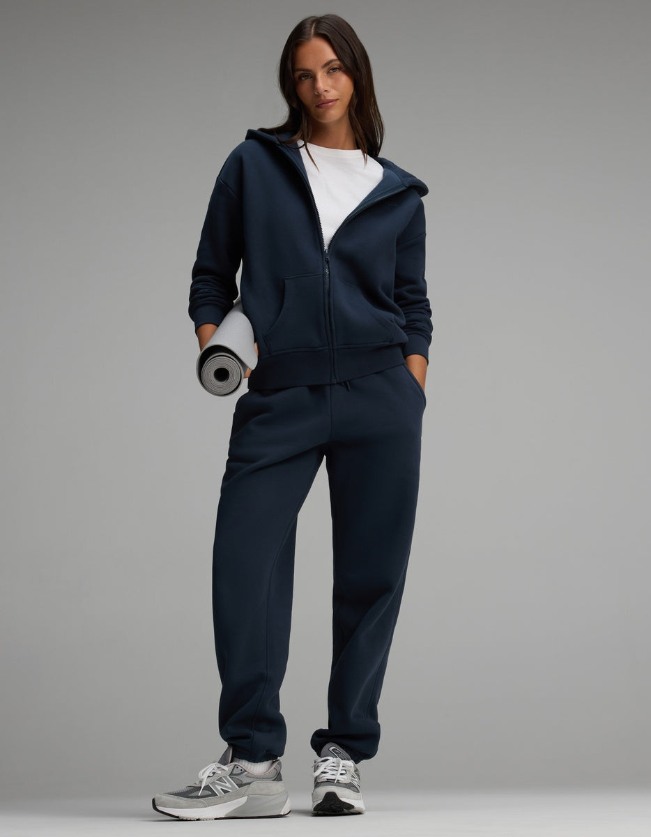 A woman wears a dark navy Women’s Cityscape Full-Zip Hoodie & Sweatpant Set and carries a rolled-up gray yoga mat.