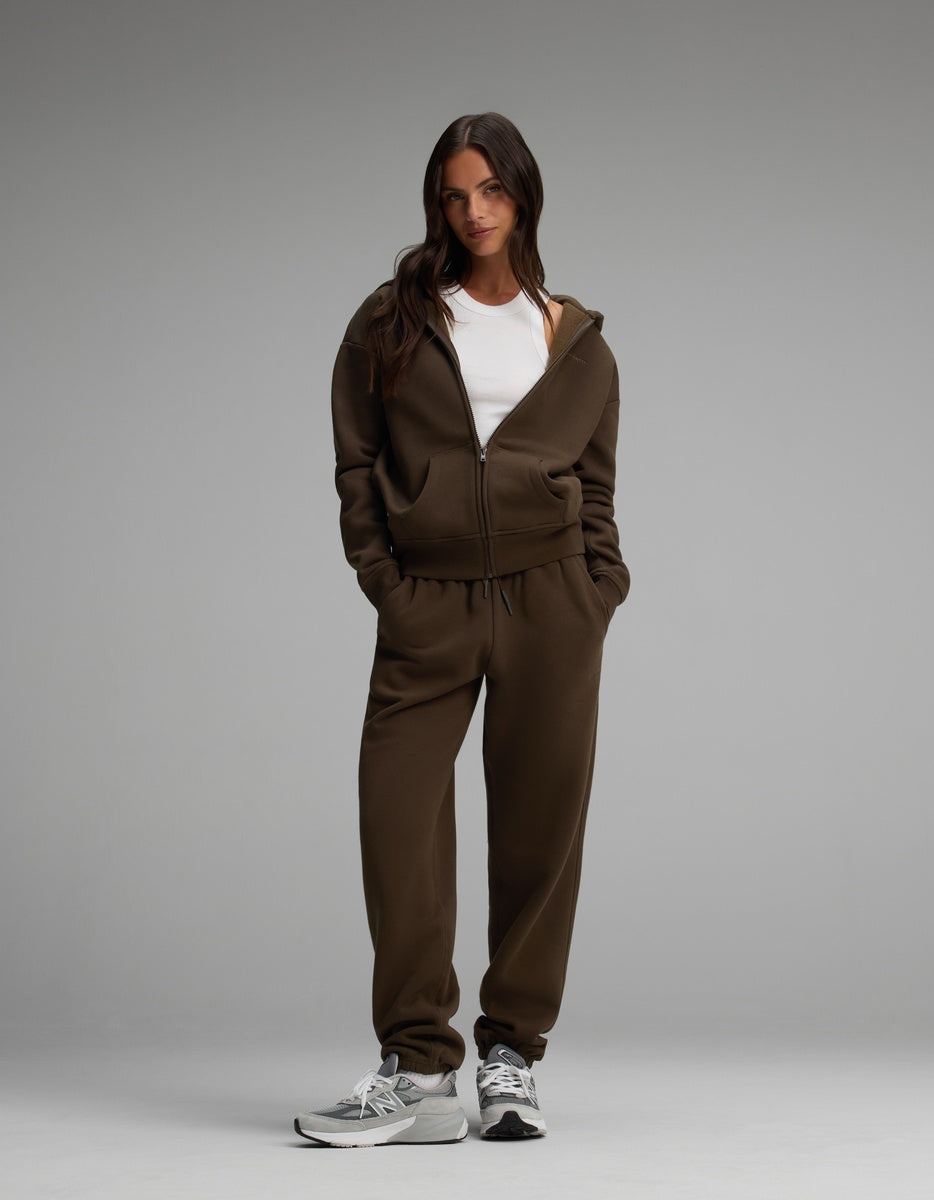 A person wears a dark brown Women’s Cityscape Full-Zip Hoodie & Sweatpant Set over a white tank top with grey sneakers.