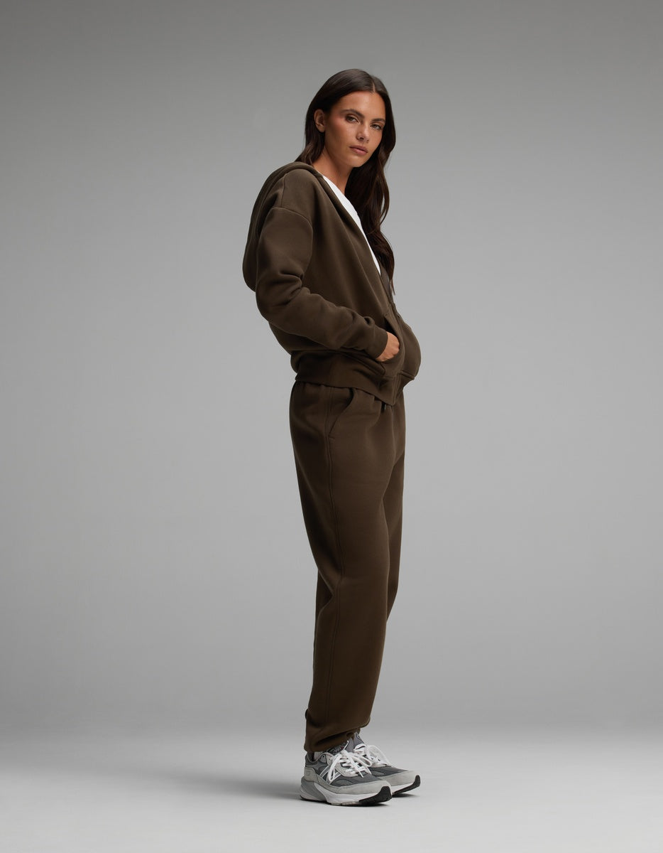 A person wearing a dark brown Women’s Cityscape Full-Zip Hoodie & Sweatpant Set with grey athletic sneakers.