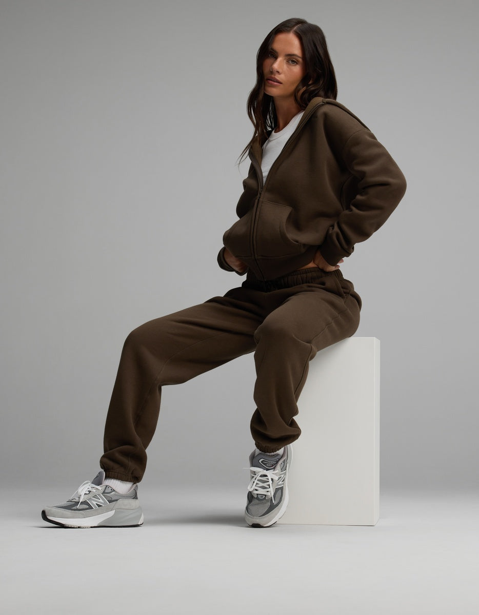 A woman wearing a brown Cozy Earth Women’s Cityscape Full-Zip Hoodie & Sweatpant Set sits on a white cube, wearing grey and white sneakers.