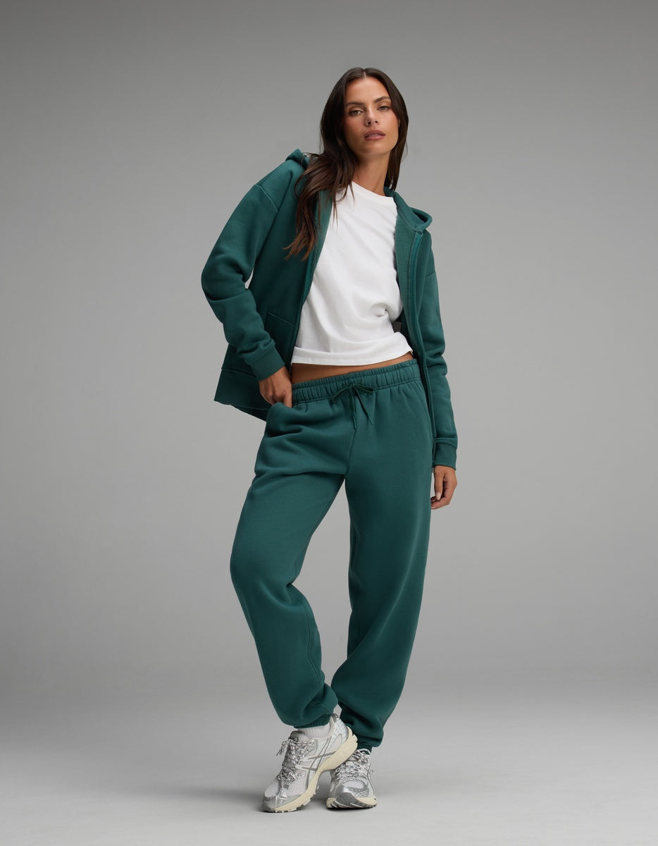 A person wears a dark teal Cozy Earth Women’s Cityscape Full-Zip Hoodie & Sweatpant Set over a white t-shirt, paired with white and silver athletic sneakers.