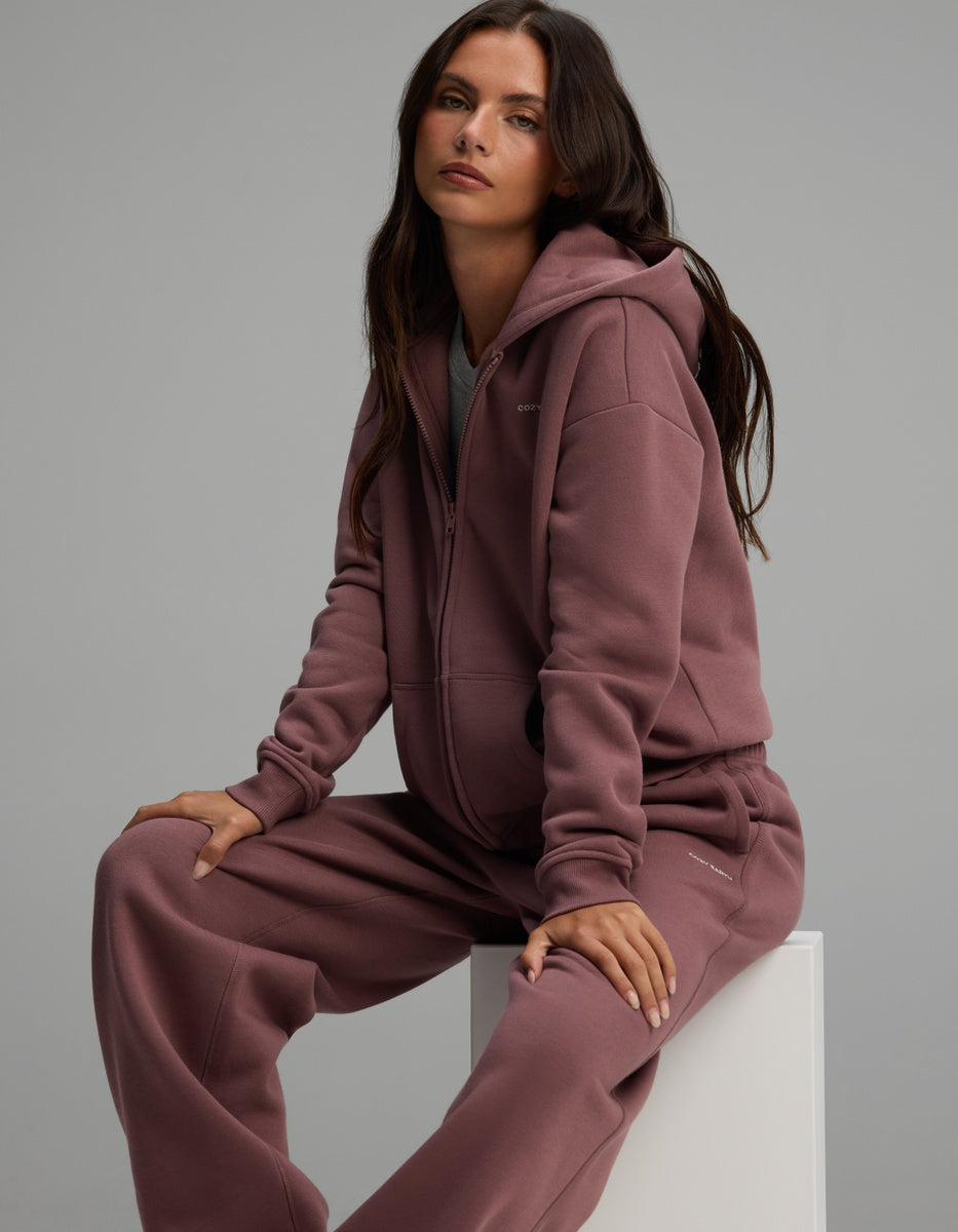 A woman wearing a mauve Women’s Cityscape Full-Zip Hoodie & Wide Leg Pant Set, sitting on a white cube.