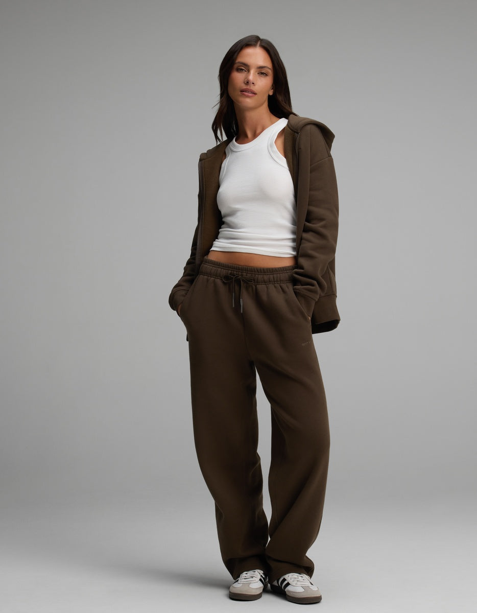 A woman wears a brown Cozy Earth Women’s Cityscape Full-Zip Hoodie & Wide Leg Pant Set over a white tank top, paired with black and white sneakers.