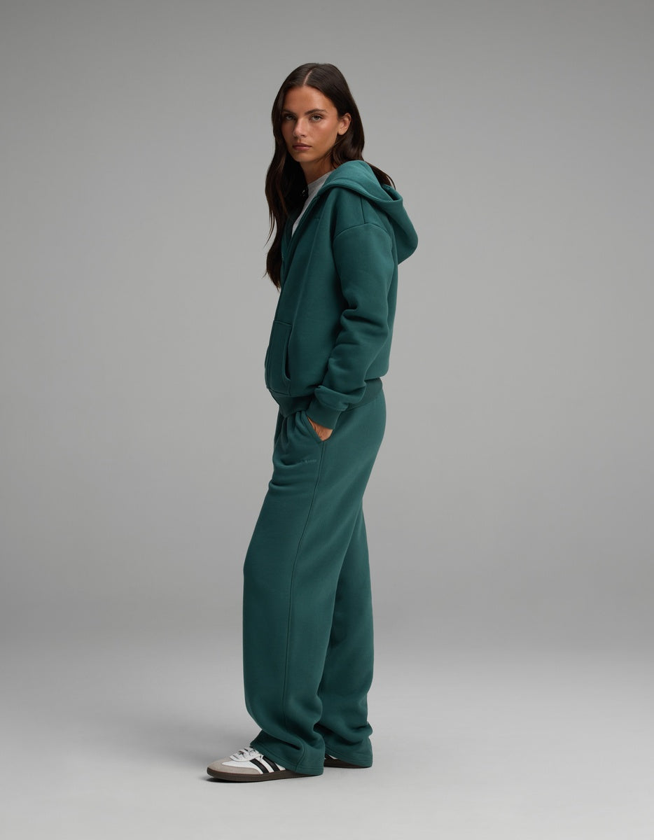 A woman standing in profile wearing a matching teal Women’s Cityscape Full-Zip Hoodie & Wide Leg Pant Set consisting of a full-zip hooded sweatshirt and wide-leg sweatpants.