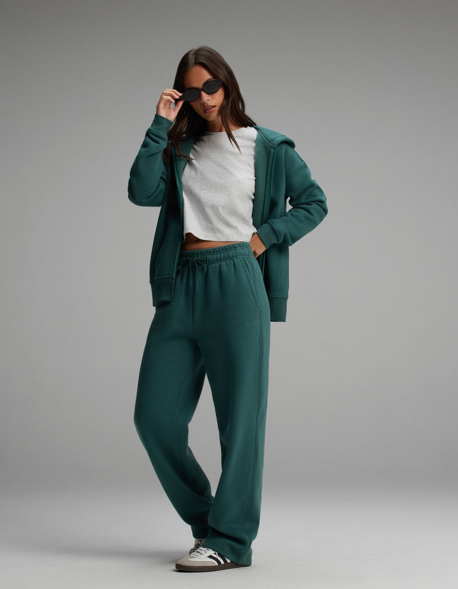 A person wearing a dark green Cozy Earth Women’s Cityscape Full-Zip Hoodie & Wide Leg Pant Set over a white t-shirt, with matching wide-leg sweatpants and sunglasses.