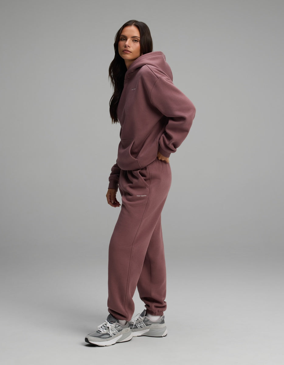 A person wearing a dusty mauve Women’s Cityscape Hoodie & Sweatpant Set with gray and white sneakers.