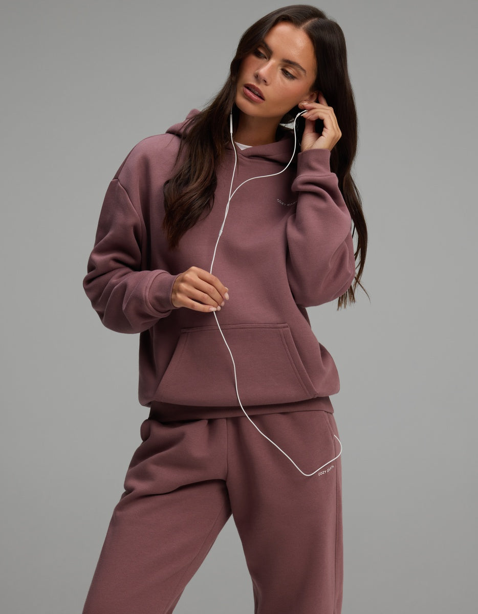 A person wearing a mauve Cozy Earth Women’s Cityscape Hoodie & Sweatpant Set, listening to wired earbuds.