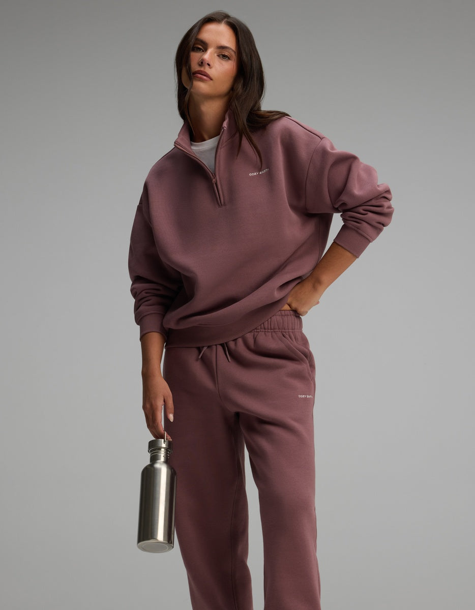 A model wearing a dusty rose Cozy Earth Women’s Cityscape Quarter Zip & Sweatpant Set, holding a metal water bottle.